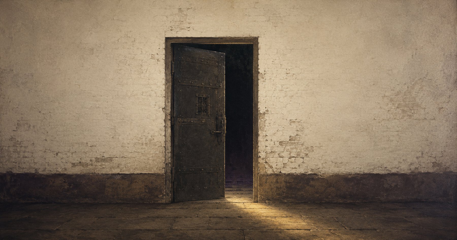 A heavy iron cell door standing ajar in a whitewashed brick wall, light falling across the stone floor at its threshold